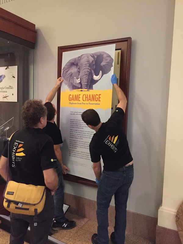 Three men in black T-shirts stand in front of a large sign with the words "Game Change" on it. The two men at the front are holding the sign up against the wall.