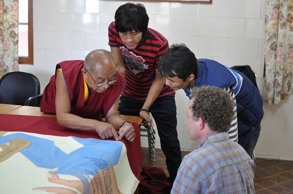 Scott watches as a monk leans over a fabric painting for the exhibit. Two other men stand and watch.