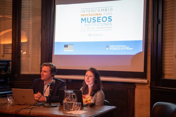 a man and a woman sit at a desk in front of a screen with the Spanish title Intercambio Profesional Para Museo e Instituciones Culturales Argentina