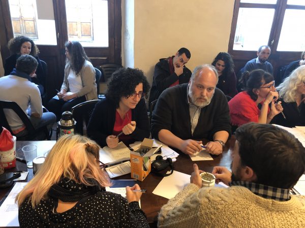 two men and two women work at a table in the foreground; other groups are working throughout the room