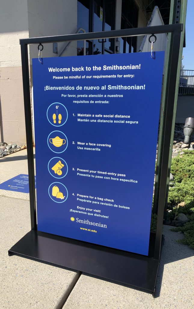 A blue graphic with white lettering and yellow icons welcomes visitors to the National Zoo. It lists the requirements for staying six feet apart, wearing a face covering, presenting a timed entry pass, and to prepare for a bag check. The graphic is in a black metal frame; the graphic can swing in the breeze to prevent the wind from knocking it over