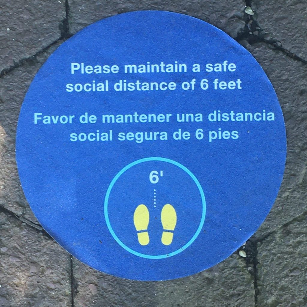 A blue circle with the words "Please maintain a safe social distance of 6 feet / Favor de mantener una distancia social segura de 6 pies" A womans toes are visible below the graphic.