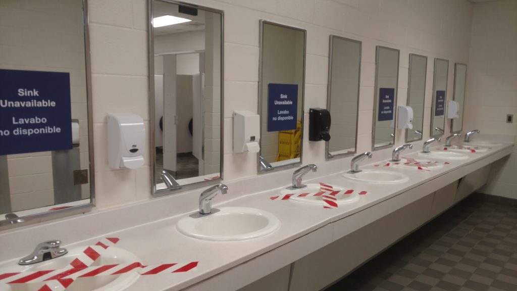 a restroom at the Smithsonian has every other sink blocked off by tape; signs on the mirrors state that the sinks are unavailable in both English and Spanish