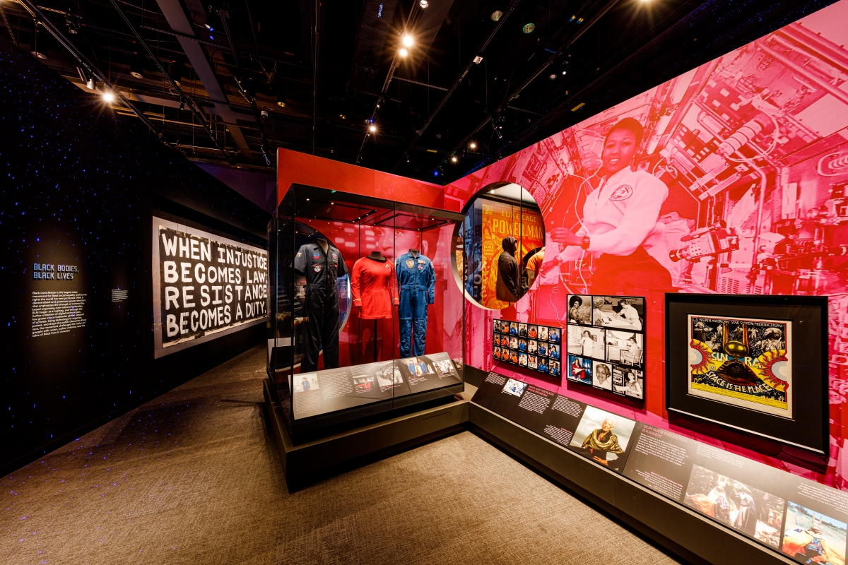 A display case features space costumes next to a pink-tinted graphic of a Black woman on a spaceship.