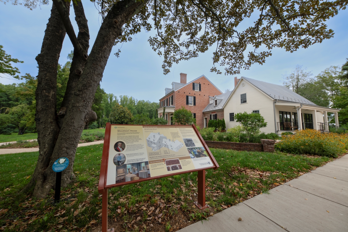 An interpretive sign featuring text and images stands in the grass in front of Woodlawn House.