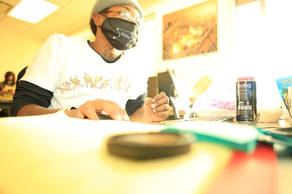 A woman wearing a face mask sits at a desk and clicks on a mouse.