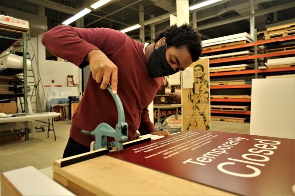 A young man wearing a face mask pushes down on a hole punch on top of a sign that reads "Temporarily Closed."